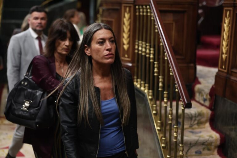 Woman with long hair in a blue top and black leather jacket walks past ornate wooden stairs with gold balusters in a formal interior setting.