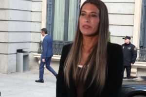 Woman with long hair in a black blazer on a city sidewalk; man in a blue suit and a security guard are visible in the background.”,