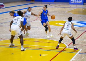 Women’s basketball game: a blue-uniform player leaps with the ball as white-uniform defenders surround her on a bright indoor court.