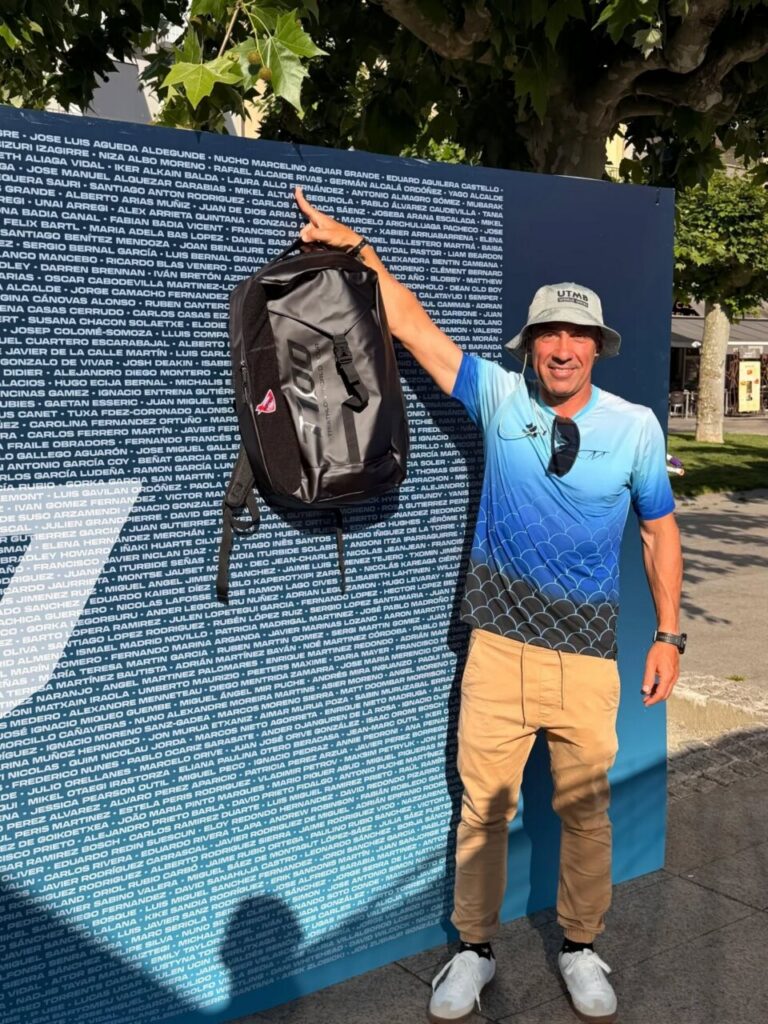 Man in a blue gradient shirt and cap holds up a black backpack beside a large banner covered in white names, outdoors in a park.
