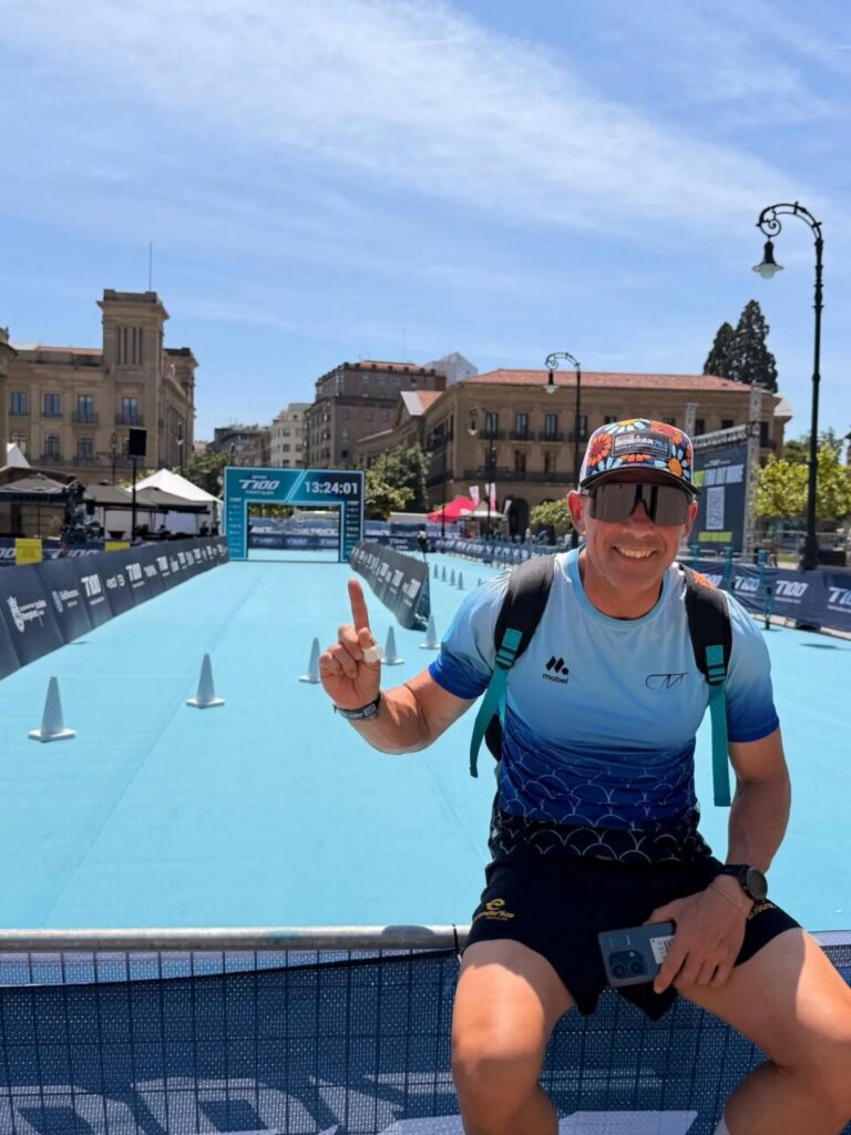 Smiling man in a blue gradient shirt sits on a barrier at a bright blue race track, pointing his index finger up with a finish-line arch in the background.
