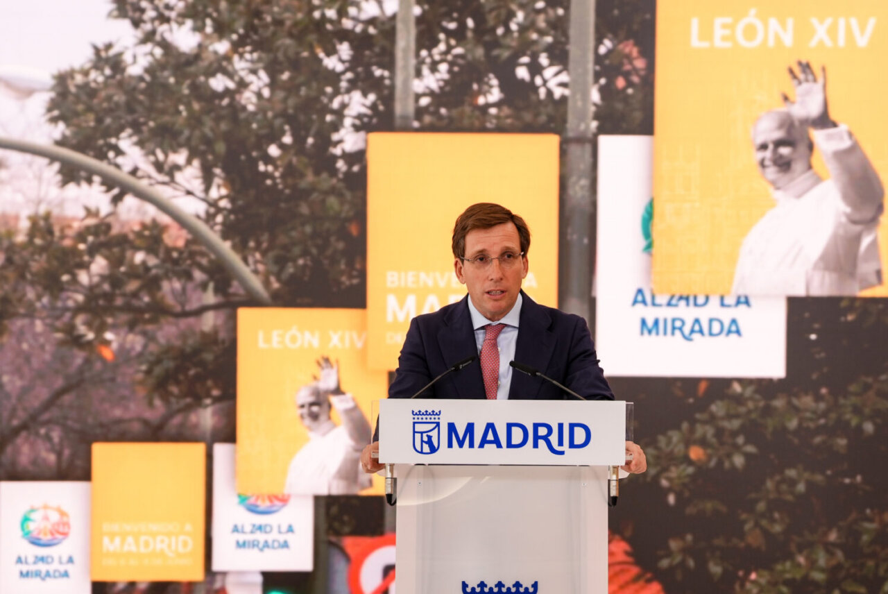 Man in a navy suit speaks at a Madrid podium with yellow event banners behind him.