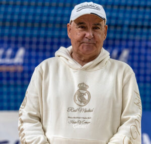 Older man in a cream Real Madrid hoodie and white cap, standing against a blue background.