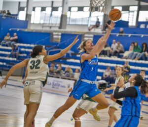 Basketball player in blue (number 3) jumps toward the hoop for a layup while defender in white (number 32) reaches up; indoor court with spectators in the background.