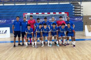 Group of futsal players and two coaches posing for a team photo in an indoor gym; blue uniforms, pink-jersey goalkeepers, and a goal behind them.