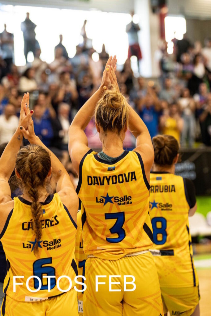 Women’s basketball players in yellow jerseys stand with hands raised in a group celebration on an indoor court, cheering crowd in the background.