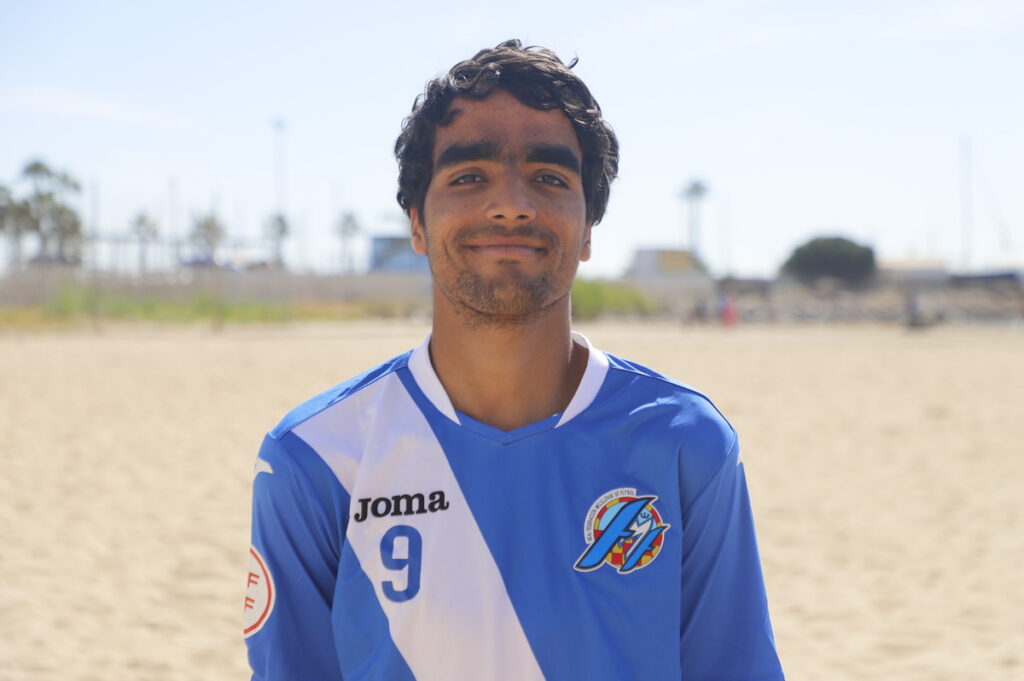 Young man standing on a sandy beach in a blue and white soccer jersey with number 9 and a crest on the chest, smiling at the camera.