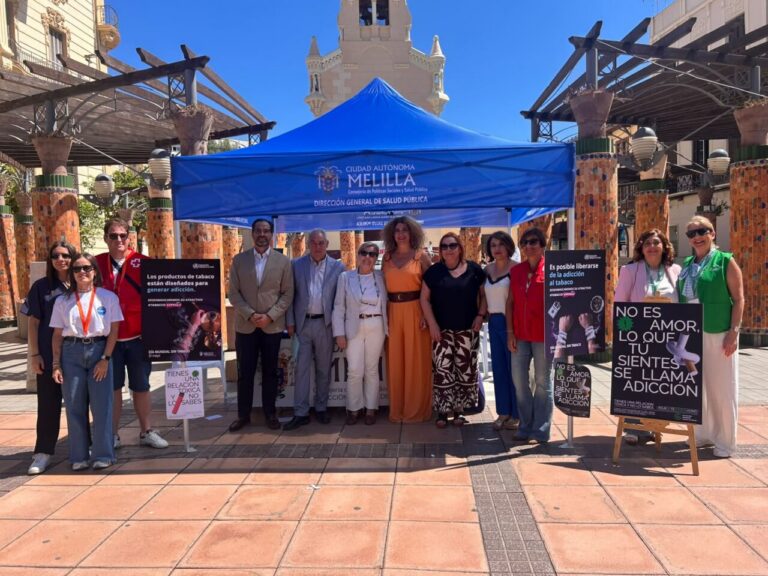 Group of people standing under a blue tent labeled 'Melilla' at a public health event in a plaza with decorative columns behind them.