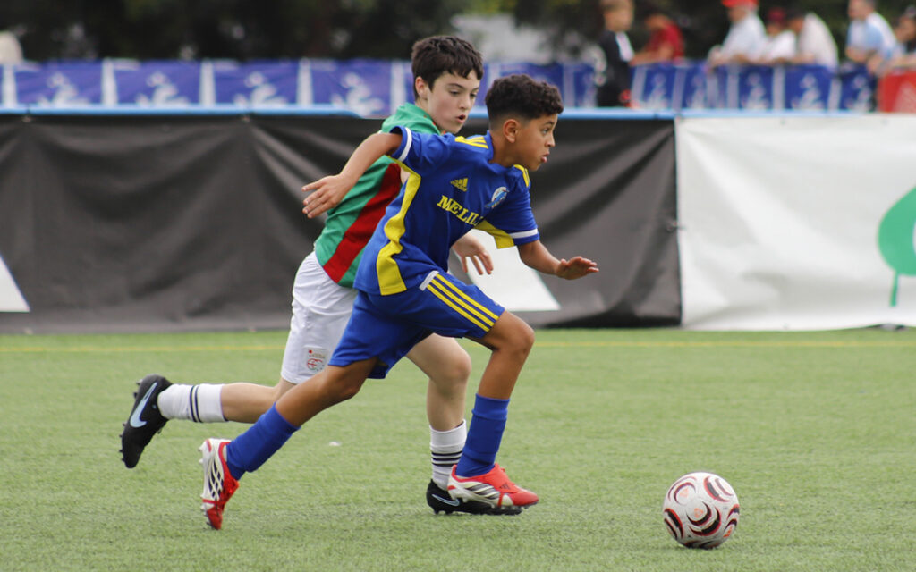 Two young soccer players chase a ball on a green field; one wears blue and yellow, the other green and red.