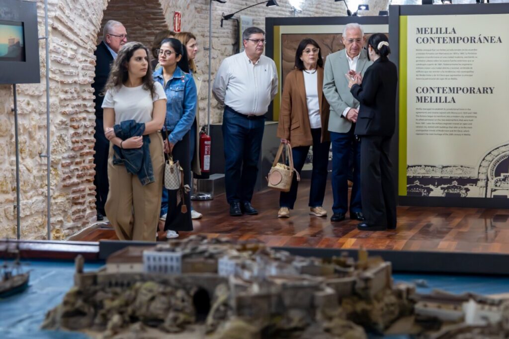 Group of visitors at a museum exhibit, standing near a large architectural diorama on a wooden floor with stone walls and text panels about Melilla Contemporánea in the background.