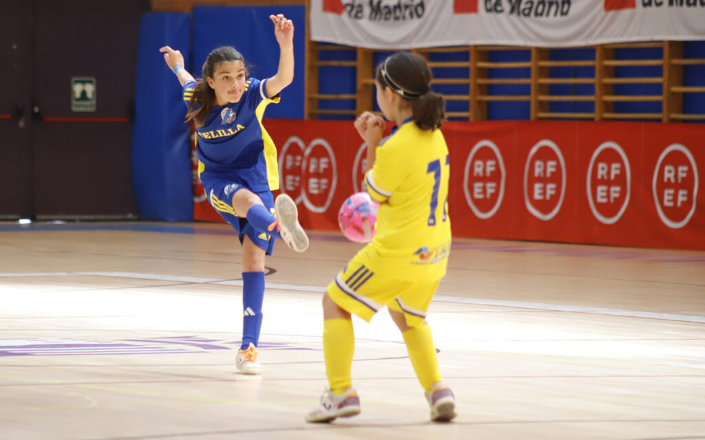 Young girl in blue kicks a pink ball in an indoor futsal court while a yellow-clad defender watches nearby.