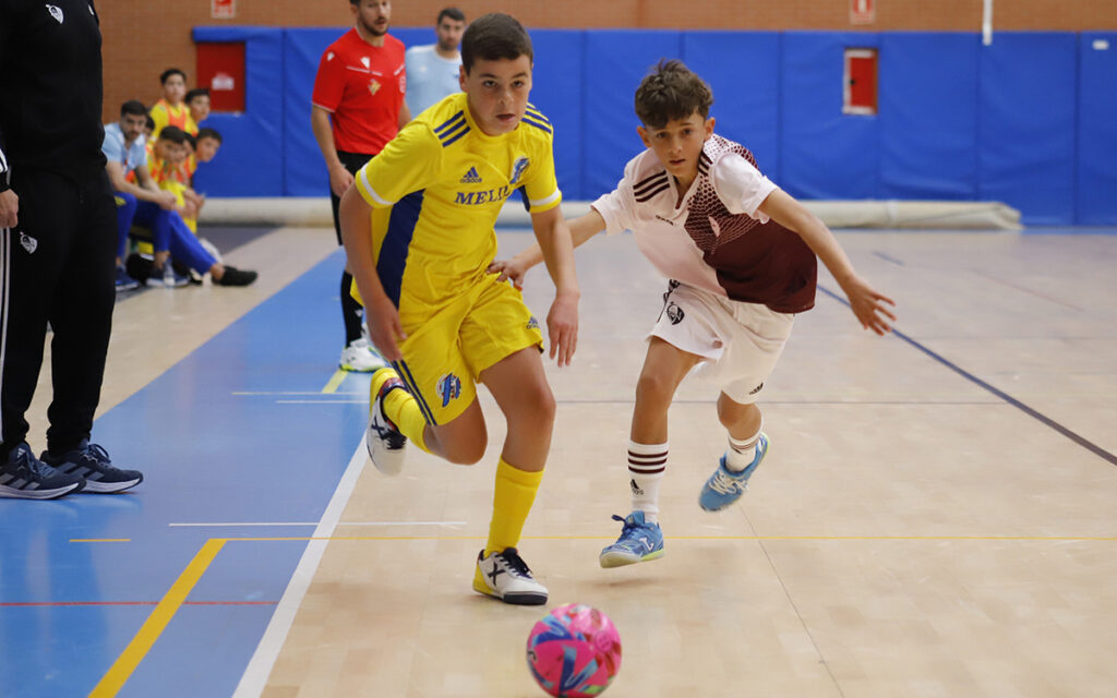 Two young futsal players race for a pink ball on an indoor court, one in a yellow kit and the other in white and maroon, with spectators along the sideline behind them.