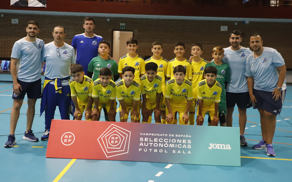 Group of young futsal players in yellow uniforms posing for a team photo with their coaches indoors beside a championship banner.
