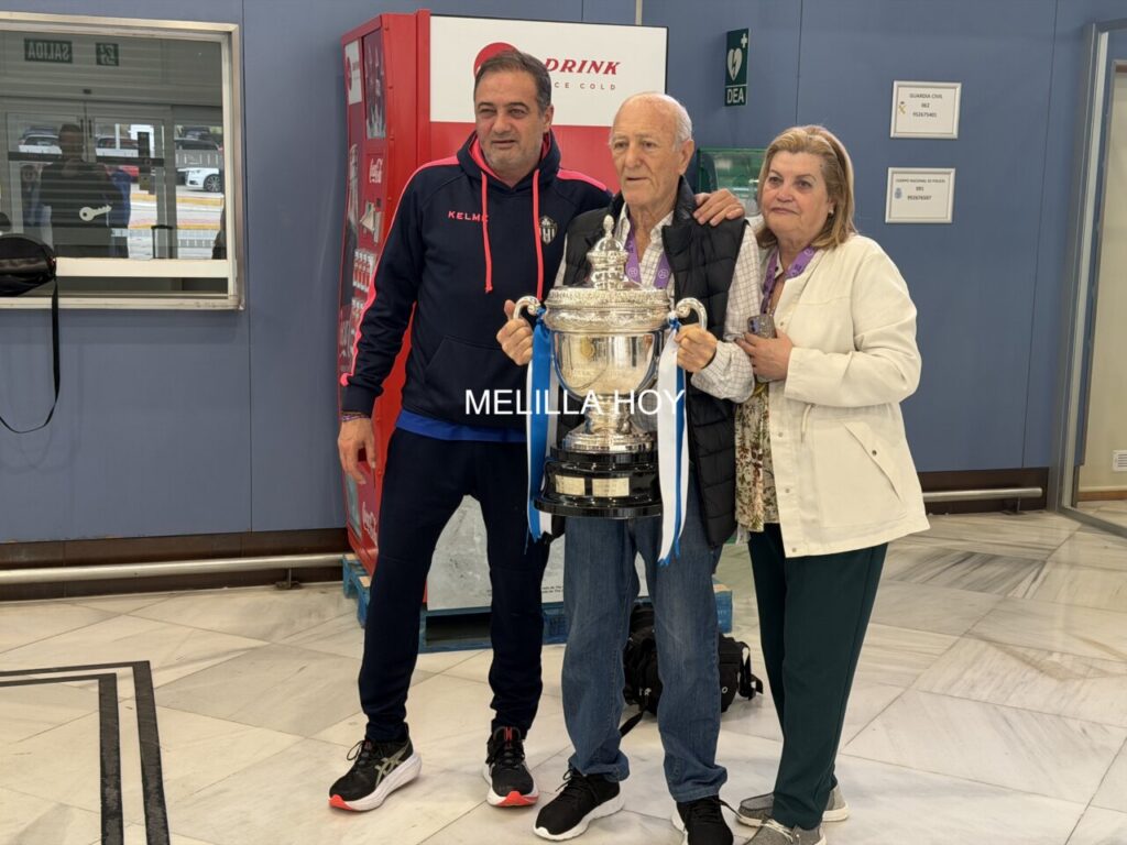 Three people posing with a large silver trophy indoors, celebrating a victory, with a vending machine in the background and a woman on the right.