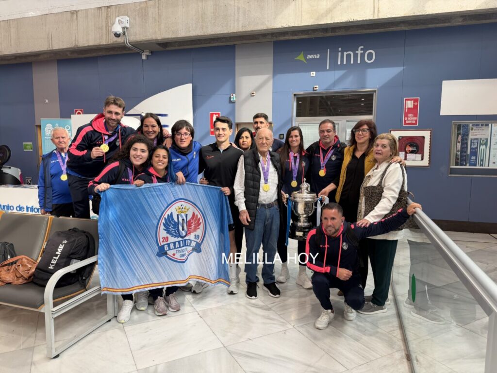 Group of athletes wearing medals posing with a banner and a large trophy inside a sports facility.
