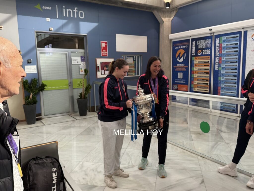 Two young women in navy jackets with pink stripes hold a large silver trophy with blue ribbons in a sports facility.