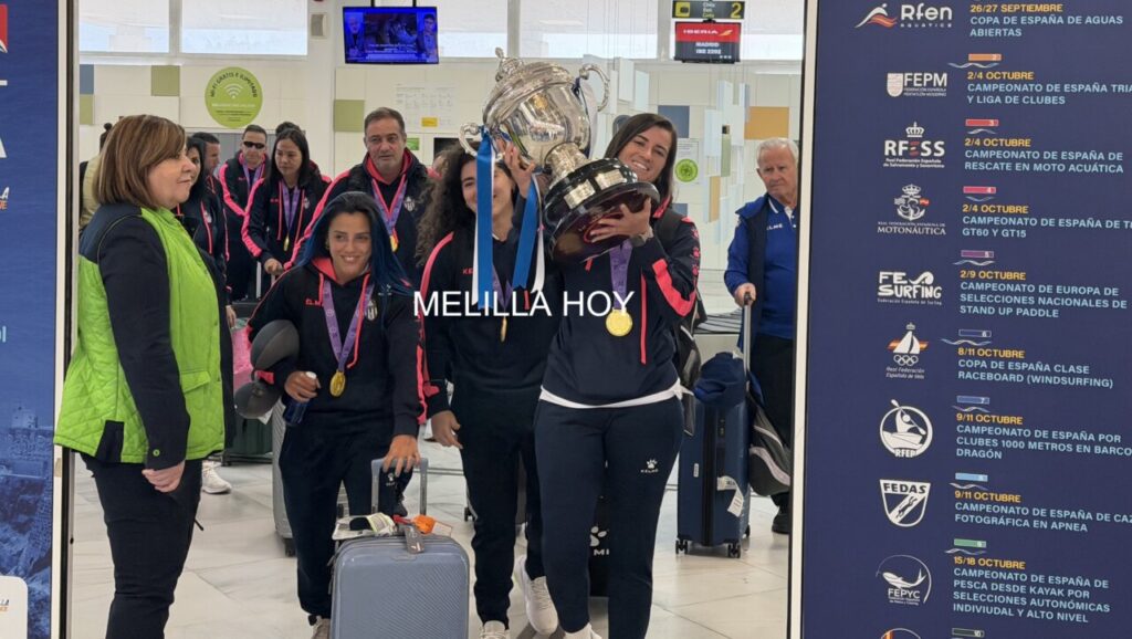 Athlete team at airport arrivals posing with a large silver trophy and gold medals, smiling smiles visible in the crowd nearby.