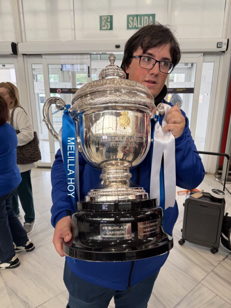 Person wearing glasses in a blue jacket holds a tall, ornate silver trophy with a blue ribbon reading 'MELILLA HOY' in a bright indoor space with an exit sign behind.