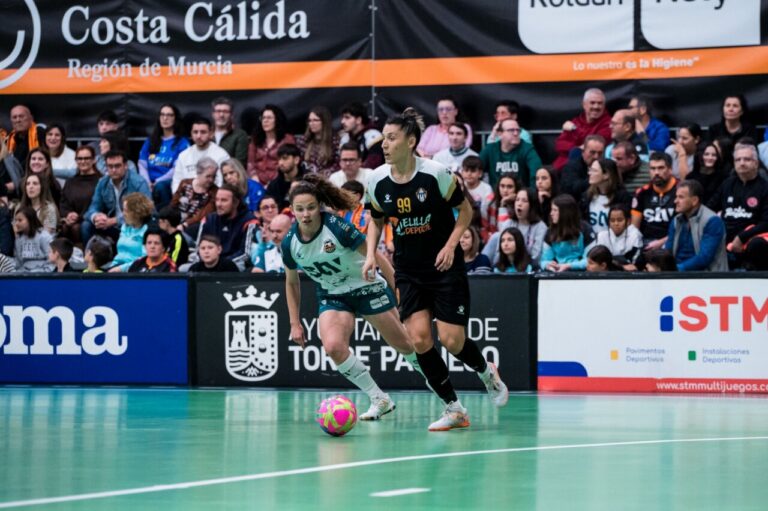 Two futsal players sprint after a bright pink ball in a packed indoor arena, with spectators watching from the stands and advertising boards along the sideline.