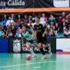 Two futsal players sprint after a bright pink ball in a packed indoor arena, with spectators watching from the stands and advertising boards along the sideline.