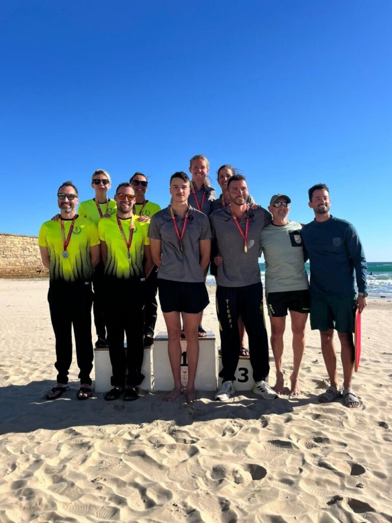 Group of athletes wearing medals posing on a beach podium with the ocean and clear blue sky in the background.