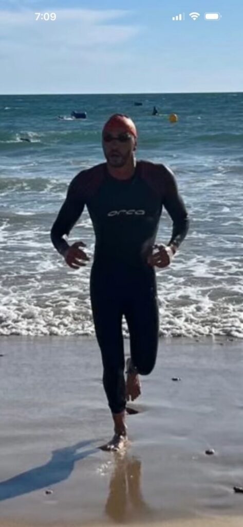 Man in a black wetsuit runs from the sea onto a sandy beach, wearing an orange swim cap and sunglasses.