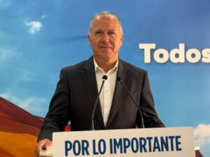 Man in a dark suit speaking at a podium with a blue backdrop reading 'POR LO IMPORTANTE' behind him.