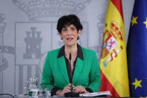 Woman in a green blazer speaks at a press conference, with a Spanish flag and EU flag in the background.
