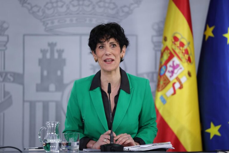 Woman in a green blazer speaks at a press conference, with a Spanish flag and EU flag in the background.