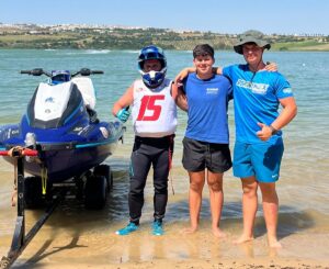 Three young men stand arm-in-arm in shallow beach water beside a blue jet ski on a trailer, smiling at the camera.