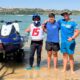 Three young men stand arm-in-arm in shallow beach water beside a blue jet ski on a trailer, smiling at the camera.