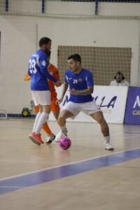 Two futsal players in blue jerseys contest a ball on an indoor court; one wears number 28, the other number 29, as a pink ball sits near their feet near the goal area in the background