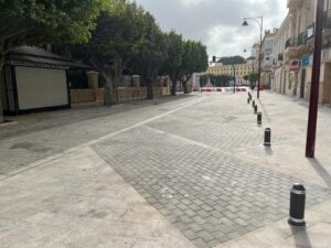 Empty pedestrian street in a European city, with trees on the left, buildings on the right, and bollards lining the walkway.
