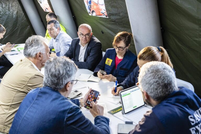 Group of professionals seated around a table in a tent, discussing with laptops, notebooks, and smartphones visible.