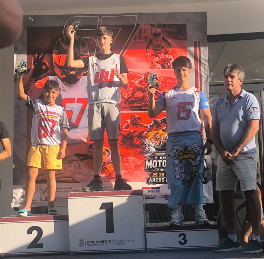Three young racers stand on a winners' podium holding trophies, with the first-place center, second on the left, and third on the right, an adult official nearby, and a large motorsport banner in the background.