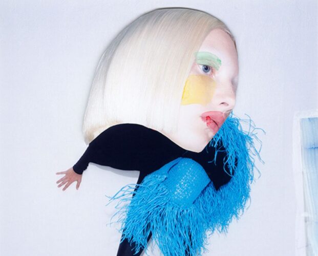 Avant-garde portrait of a person with an oversized blonde bob, wearing a black outfit with blue feathers and bold face makeup (green eyebrow, yellow cheek, red lips).
