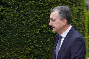 Side profile of a middle-aged man in a dark navy suit and lavender tie, standing in front of a green hedge backdrop.