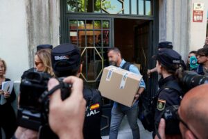 Man carrying a large taped cardboard box toward a doorway, flanked by Policía Nacional officers and reporters outside a building.