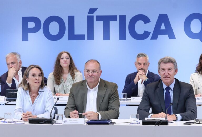 Panel of six professionals seated at a long table during a press conference with a blue backdrop reading 'POLÍTICA', microphones and nameplates in front of them