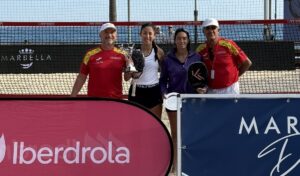 Four players pose with trophies on a beach tennis court, two men in red shirts on ends and two women in center holding paddles and medals.