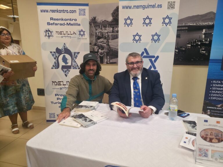 Two men sit at a table signing books at a promotional booth, flanked by banners with logos; a woman stands left holding a cardboard box.