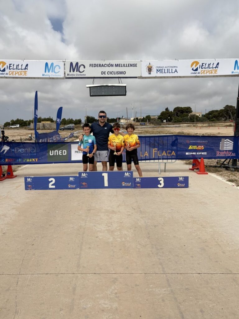 Man and three young cyclists stand on the 1st place podium at a cycling race, posing for a photo with banners and finish line overhead in a sunny outdoor venue.