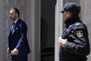 Man in a dark blue suit steps out of a doorway while a security officer in a dark uniform stands nearby outside the entrance.