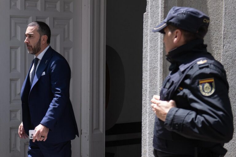 Man in a dark blue suit steps out of a doorway while a security officer in a dark uniform stands nearby outside the entrance.