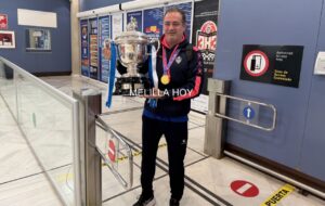 Man in athletic wear with a gold medal, holding a large silver trophy with blue ribbons in a sports facility.