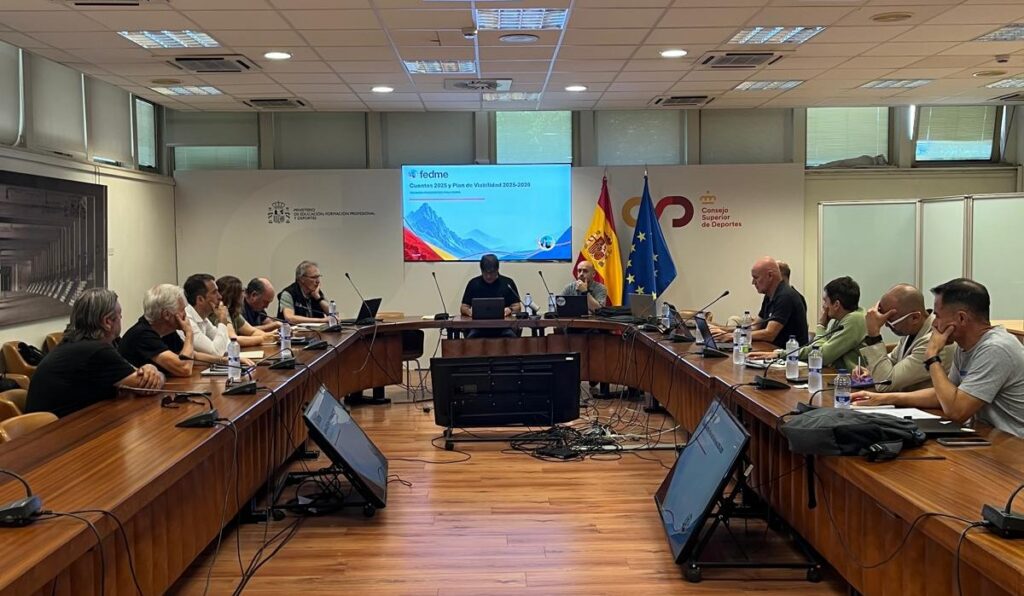 Group of people seated around a U-shaped conference table in a meeting room; presentation screen and Spain/EU flags behind.