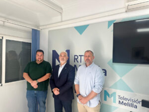 Three men stand together in a TV studio with a light blue geometric logo wall and a mounted screen behind them.