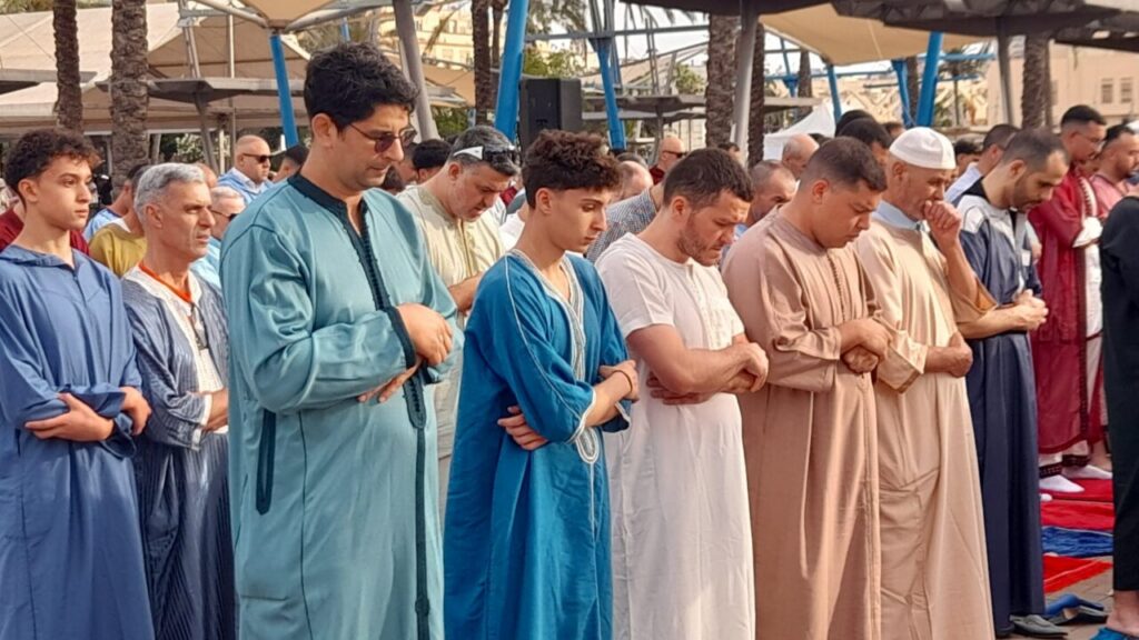 Group of men in traditional robes standing in prayer with folded arms in an outdoor gathering.