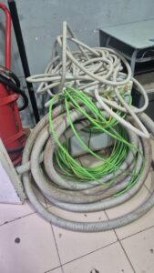 Messy pile of grey corrugated hoses and bright green cables coiled on a tiled workshop floor next to a cabinet and equipment.