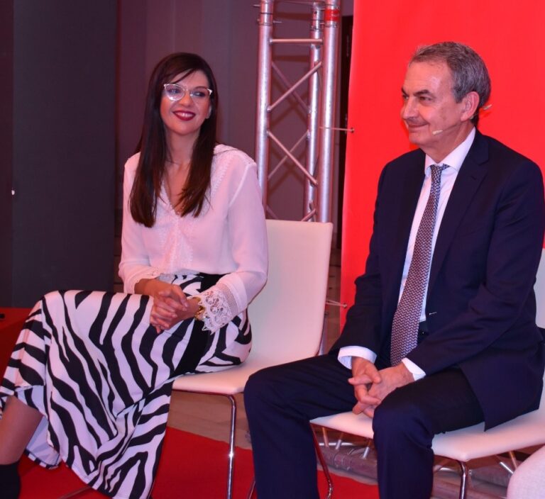 Two speakers seated on stage, woman in a white blouse and zebra-print skirt smiling, man in a dark suit with a patterned tie listening.